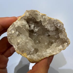 Peut inclure: Une géode, formation rocheuse creuse, est tenue. L'extérieur est de couleur beige clair, tandis que l'intérieur révèle un amas de structures cristallines claires. Les cristaux scintillent, créant un contraste visuellement attrayant.