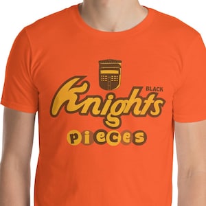 Puede incluir: Camiseta naranja de manga corta con el texto "Knights Pieces" en un diseño amarillo y marrón. La palabra "Black" está encima de la palabra "Knights". La camiseta está hecha de un material suave.
