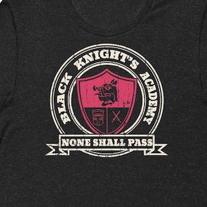 Peut inclure: T-shirt gris foncé avec un motif vieilli. Le motif comprend un emblème circulaire avec le texte "BLACK KNIGHT'S ACADEMY" et un écusson noir et rose. En dessous, le texte indique "NONE SHALL PASS."