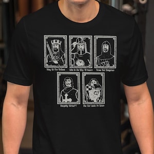 Puede incluir: Una camiseta negra de manga corta con un estampado gráfico blanco de cinco ilustraciones de personajes de estilo medieval dispuestas en una cuadrícula, cada una acompañada de pequeñas etiquetas de texto que incluyen King of the Britons.