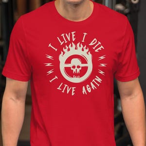 Puede incluir: Camiseta roja con el texto blanco "I LIVE I DIE I WANT I LIVE AGAIN" que rodea un diseño de calavera y llamas. La camiseta de manga corta parece estar hecha de un material suave.