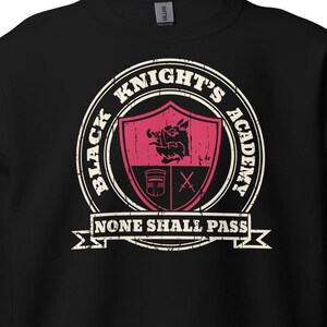 Peut inclure: Sweat-shirt noir avec un motif vieilli. Le motif présente un écusson avec les mots "Black Knight's Academy" dans un design circulaire. En dessous, une bannière indique "None Shall Pass".