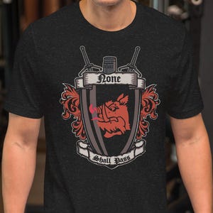 May include: Dark gray t-shirt featuring a crest design with a red boar, banner with the word "None", and the phrase "Shall Pass". The crest is framed by decorative elements and crossed swords.