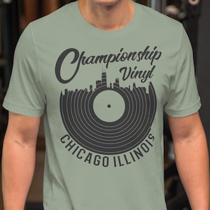 May include: Sage green t-shirt with a black graphic of a vinyl record and the Chicago skyline. The text on the shirt reads "Championship Vinyl Chicago Illinois". The shirt is short-sleeved and has a crew neck.