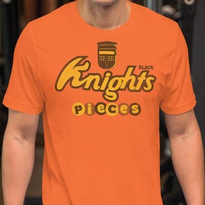 May include: Orange t-shirt with the words "Knights" and "Pieces" in a brown, stylized font. The word "Black" is above the word "Knights". The shirt is short-sleeved and made of a soft material.