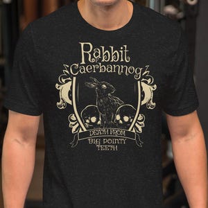 Puede incluir: Camiseta gris oscuro con una ilustración de conejo y el texto "Rabbit Caerbannog" arriba y "Death from Big Pointy Teeth" abajo. El diseño incluye calaveras y elementos decorativos.