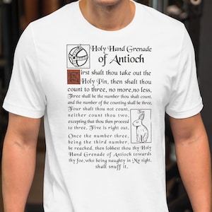 Peut inclure: T-shirt blanc avec l'inscription "Holy Hand Grenade of Antioch" et un graphisme détaillé. Le motif comprend une bordure décorative, une illustration de Holy Pin et un lapin. Le t-shirt est à col rond et à manches courtes.