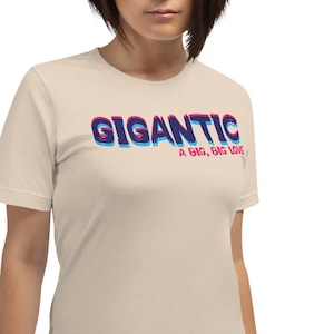 Puede incluir: Camiseta beige con la palabra "GIGANTIC" en una fuente en capas roja y azul. Debajo, la frase "A BIG, BIG LOVE". La camiseta es de manga corta y tiene cuello redondo.