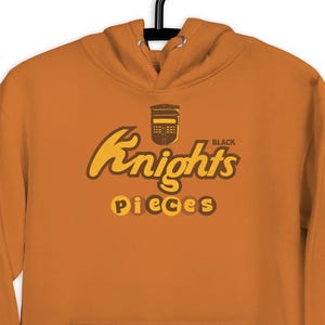 May include: An orange hoodie with a front pocket and a drawstring hood. The hoodie features the text "Knights Pieces" in a brown and yellow design. The hoodie is hanging on a black hanger.