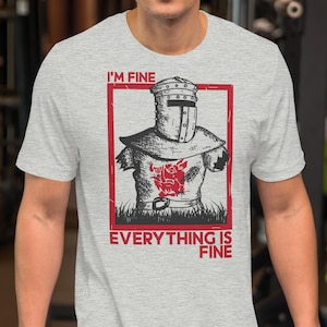 Peut inclure: T-shirt gris clair avec un motif de chevalier dans un cadre rouge. Le texte "I'M FINE" est en haut et "EVERYTHING IS FINE" en bas. Le motif du chevalier est noir et rouge.