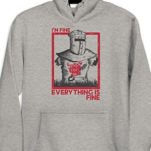 Peut inclure: Un sweat à capuche gris chiné avec un motif graphique. Le motif représente un chevalier en armure avec le texte "I'M FINE" au-dessus et "EVERYTHING IS FINE" en dessous, le tout dans un rectangle rouge. Le design est de style vintage.