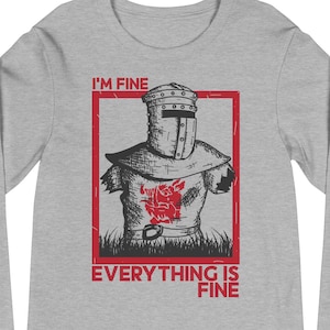 Peut inclure: Un t-shirt gris à manches longues avec un dessin rouge et noir d'un chevalier en armure. Le texte "I'M FINE" est au-dessus et "EVERYTHING IS FINE" en dessous. L'illustration du chevalier est dans un rectangle rouge.