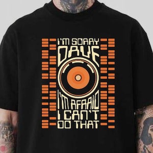 Puede incluir: Camiseta negra con el texto "I'M SORRY DAVE I'M AFRAID I CAN'T DO THAT" en naranja y blanco. El diseño incluye un gráfico de disco y formas rectangulares.