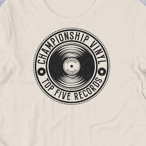 May include: Cream-colored t-shirt featuring a black graphic of a vinyl record with the text "CHAMPIONSHIP VINYL TOP FIVE RECORDS" in a circular design. The shirt is made of a soft material and is displayed on a neutral surface with other accessories.