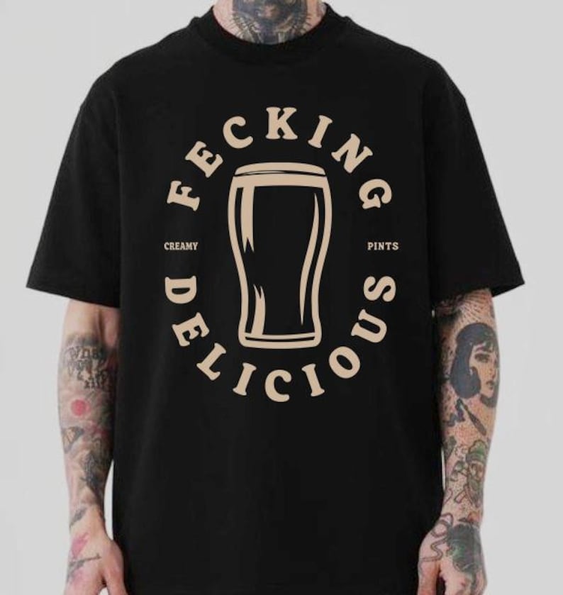 May include: Black t-shirt with a circular graphic featuring a pint glass illustration. The text around the glass reads "Feckin' Delicious Creamy Pints" in a beige color. The shirt has short sleeves and a crew neck.