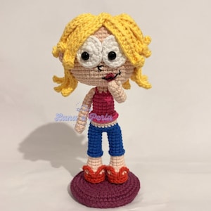 Può includere: Bambola all'uncinetto fatta a mano con capelli biondi, un top rosa, pantaloni blu e scarpe rosse. La bambola è in piedi su una base viola. La bambola ha grandi occhi bianchi e un'espressione sorridente.