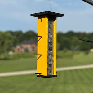 May include: A yellow bird feeder with a black top and bottom. The feeder has a rectangular shape with a mesh panel on one side and three small perches. The background shows a green lawn and trees.