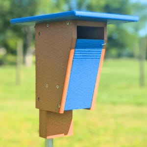 May include: A brown and blue birdhouse with an orange accent. The birdhouse has a blue roof and a blue entrance ramp. The birdhouse is mounted on a metal pole and is set against a blurred green background.