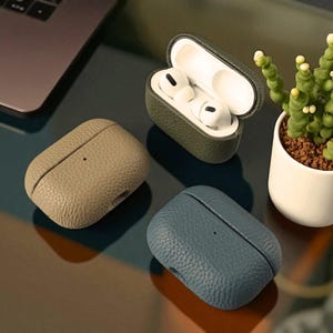 May include: Three leather AirPods Pro cases in beige, green, and blue. One case is open, revealing white earbuds. The cases have a textured surface and a small hole for charging. A small potted plant is in the background.