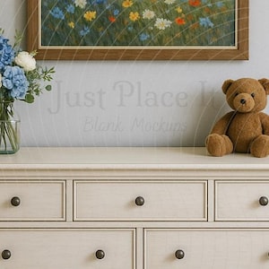 Könnte beinhalten: Eine cremefarbene Kommode mit sieben Schubladen und dunklen Knöpfen. Ein gerahmtes Gemälde eines Wildblumenfeldes hängt über der Kommode. Ein brauner Teddybär sitzt auf der Kommode, neben einer Blumenvase.