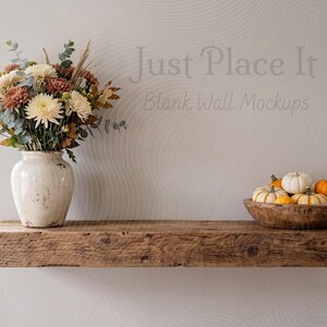 May include: A rustic wooden floating shelf mounted on a wall, featuring a cream-coloured ceramic vase filled with autumn flowers and a wooden bowl containing small white and orange pumpkins. The wall displays the text Just Place It Blank Wall Mockups.