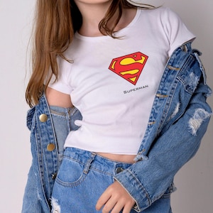 K&ouml;nnte beinhalten: Wei&szlig;es T-Shirt mit dem Superman-Logo in Rot und Gelb und dem Wort "Superman" darunter. Dazu eine Jeansjacke und blaue Jeans, f&uuml;r einen l&auml;ssigen, allt&auml;glichen Look.