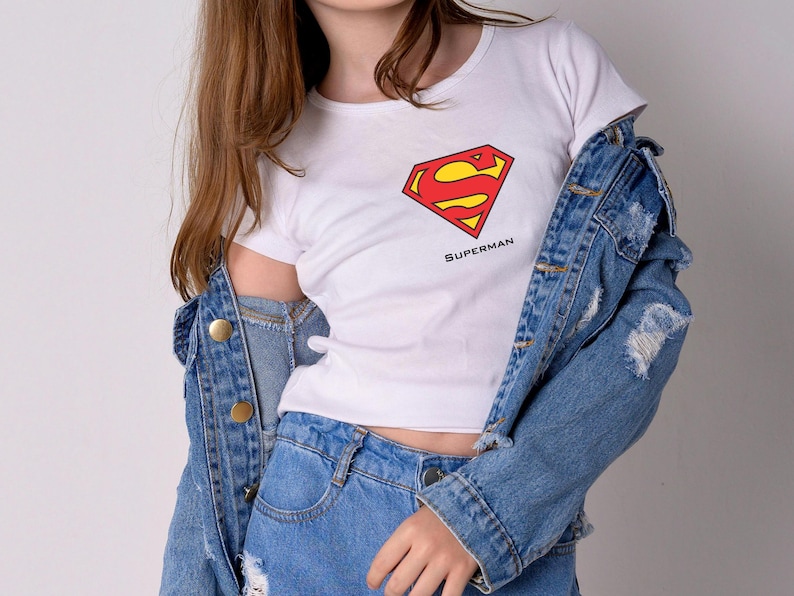 K&ouml;nnte beinhalten: Wei&szlig;es T-Shirt mit dem Superman-Logo in Rot und Gelb und dem Wort "Superman" darunter. Dazu eine Jeansjacke und blaue Jeans, f&uuml;r einen l&auml;ssigen, allt&auml;glichen Look.