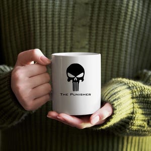 K&ouml;nnte beinhalten: Wei&szlig;e Keramik-Tasse mit einem schwarzen Sch&auml;del-Design und dem Text "THE PUNISHER". Die Tasse wird von einer Person gehalten, die einen gr&uuml;nen Pullover tr&auml;gt. Eine Kaffeetasse. Fassungsverm&ouml;gen ca. 300 ml.