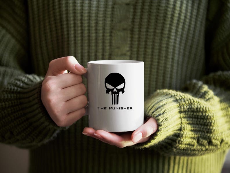 K&ouml;nnte beinhalten: Wei&szlig;e Keramik-Tasse mit einem schwarzen Sch&auml;del-Design und dem Text "THE PUNISHER". Die Tasse wird von einer Person gehalten, die einen gr&uuml;nen Pullover tr&auml;gt. Eine Kaffeetasse. Fassungsverm&ouml;gen ca. 300 ml.
