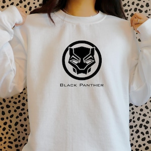 K&ouml;nnte beinhalten: Wei&szlig;es Sweatshirt mit einem schwarzen Black Panther-Logo in einem kreisf&ouml;rmigen Design. Der Text "BLACK PANTHER" ist unter dem Logo aufgedruckt. Das Sweatshirt ist aus einem weichen Material gefertigt.