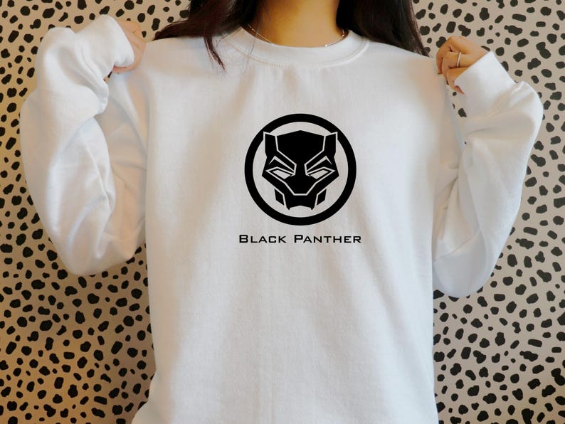 K&ouml;nnte beinhalten: Wei&szlig;es Sweatshirt mit einem schwarzen Black Panther-Logo in einem kreisf&ouml;rmigen Design. Der Text "BLACK PANTHER" ist unter dem Logo aufgedruckt. Das Sweatshirt ist aus einem weichen Material gefertigt.