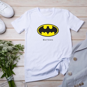 K&ouml;nnte beinhalten: Wei&szlig;es T-Shirt mit dem Batman-Logo in Schwarz und Gelb und dem Wort "BATMAN" darunter. Das Shirt wird mit wei&szlig;en Turnschuhen, einem hellgrauen Blazer und einem Strau&szlig; wei&szlig;er Blumen gezeigt.