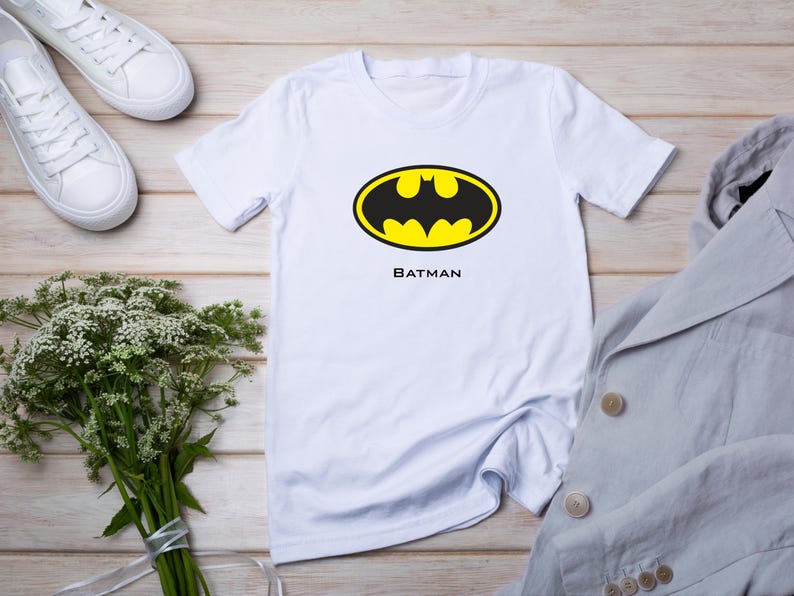 K&ouml;nnte beinhalten: Wei&szlig;es T-Shirt mit dem Batman-Logo in Schwarz und Gelb und dem Wort "BATMAN" darunter. Das Shirt wird mit wei&szlig;en Turnschuhen, einem hellgrauen Blazer und einem Strau&szlig; wei&szlig;er Blumen gezeigt.