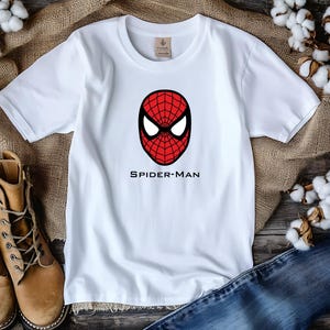 K&ouml;nnte beinhalten: Wei&szlig;es T-Shirt mit einem rot-schwarzen Spider-Man-Masken-Motiv und dem Text "SPIDER-MAN" darunter. Das T-Shirt wird auf einer Holzoberfl&auml;che mit Jeans und braunen Stiefeln pr&auml;sentiert. Baumwollbl&uuml;ten im Hintergrund.