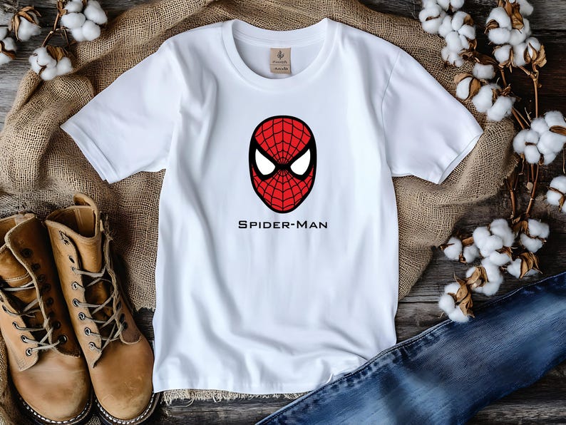 K&ouml;nnte beinhalten: Wei&szlig;es T-Shirt mit einem rot-schwarzen Spider-Man-Masken-Motiv und dem Text "SPIDER-MAN" darunter. Das T-Shirt wird auf einer Holzoberfl&auml;che mit Jeans und braunen Stiefeln pr&auml;sentiert. Baumwollbl&uuml;ten im Hintergrund.