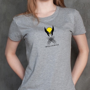 K&ouml;nnte beinhalten: Ein graues T-Shirt mit dem Wolverine-Logo. Das Logo zeigt einen gelben Kreis mit schwarzen Akzenten, ein schwarzes Maskendesign und drei Krallen. Das Wort "WOLVERINE" ist unter der Grafik aufgedruckt.
