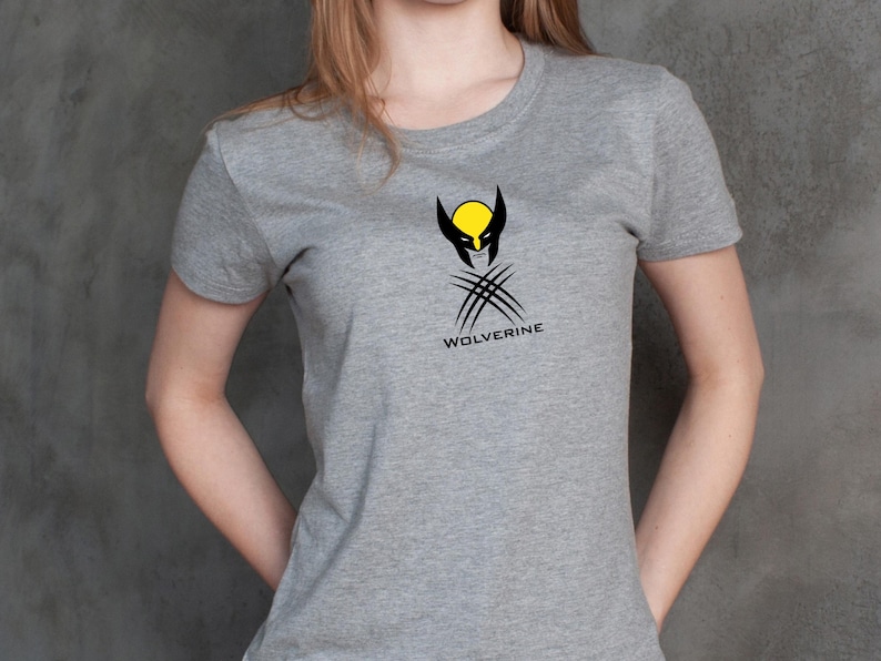 K&ouml;nnte beinhalten: Ein graues T-Shirt mit dem Wolverine-Logo. Das Logo zeigt einen gelben Kreis mit schwarzen Akzenten, ein schwarzes Maskendesign und drei Krallen. Das Wort "WOLVERINE" ist unter der Grafik aufgedruckt.