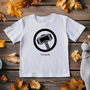 K&ouml;nnte beinhalten: Wei&szlig;es T-Shirt mit einer schwarzen Grafik von Thors Hammer in einem schwarzen Kreis. Darunter steht das Wort "THOR". Das Shirt liegt auf einer Holzoberfl&auml;che mit Herbstbl&auml;ttern.