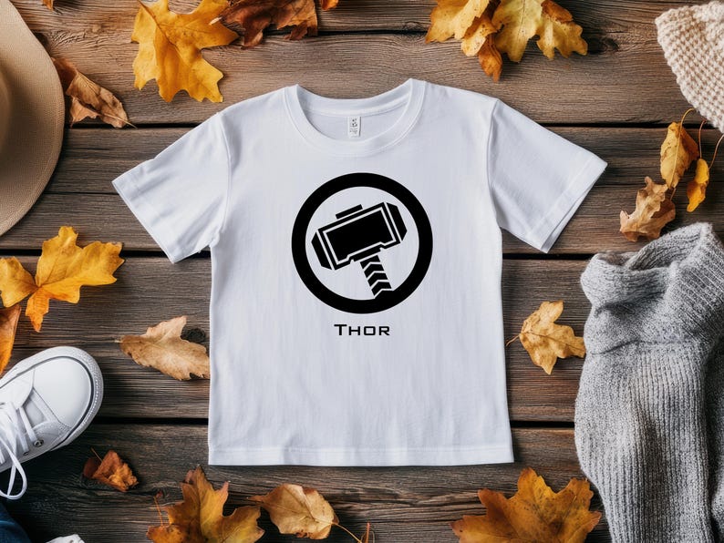 K&ouml;nnte beinhalten: Wei&szlig;es T-Shirt mit einer schwarzen Grafik von Thors Hammer in einem schwarzen Kreis. Darunter steht das Wort "THOR". Das Shirt liegt auf einer Holzoberfl&auml;che mit Herbstbl&auml;ttern.