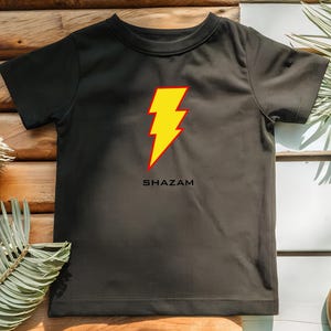 K&ouml;nnte beinhalten: Schwarzes T-Shirt mit einem gelb-roten Blitz-Design und dem Wort "SHAZAM" darunter. Das kurz&auml;rmelige Shirt ist aus einem weichen Material. Ideal f&uuml;r Superhelden-Fans.