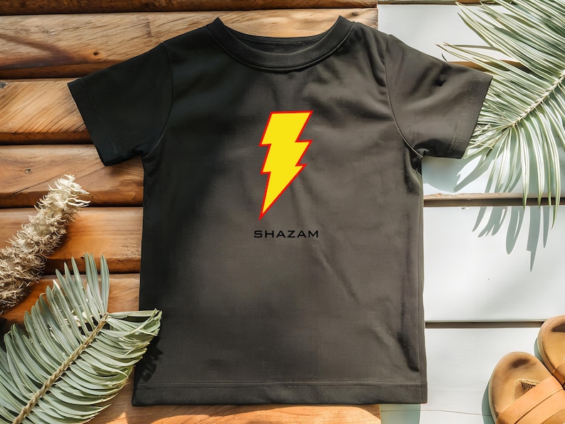 K&ouml;nnte beinhalten: Schwarzes T-Shirt mit einem gelb-roten Blitz-Design und dem Wort "SHAZAM" darunter. Das kurz&auml;rmelige Shirt ist aus einem weichen Material. Ideal f&uuml;r Superhelden-Fans.