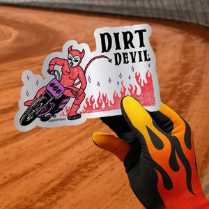 May include: A sticker featuring a red devil riding a motorcycle with the number 666. The sticker has the words "DIRT DEVIL" in black text, with flames and purple stars. The sticker is held by a hand wearing a black, orange, and yellow glove.