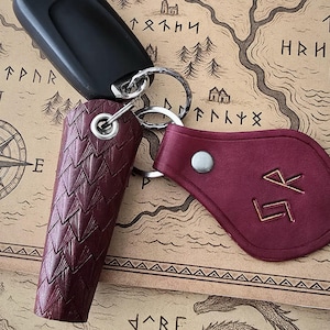 May include: Keys with a black remote, a burgundy leather key holder with a textured pattern, and a burgundy leather keychain with gold runes. The keys are on a map.