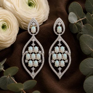 May include: A pair of silver-toned drop earrings featuring a marquise-shaped frame encrusted with small clear stones. Each earring contains a central cluster of nine oval-shaped white opalescent cabochons and one additional stone at the top.