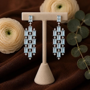 May include: A pair of long drop earrings featuring a vertical arrangement of rectangular light blue gemstones and small round clear stones set in a silver-toned metal frame. The design consists of multiple linked segments creating a linear pattern.