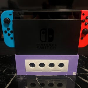 Könnte beinhalten: Eine Nintendo Switch-Konsole mit blauen und roten Joy-Con-Controllern. Die Konsole ist in einer lila GameCube-Basis mit vier kreisförmigen Anschlüssen angedockt. Das Nintendo Switch-Logo ist sichtbar.