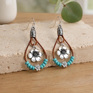 May include: A pair of dangle earrings featuring a brown leather teardrop loop, a silver-toned metal flower charm, and a row of small turquoise-colored beads. The earrings are finished with silver-toned metal hooks and decorative bead caps.
