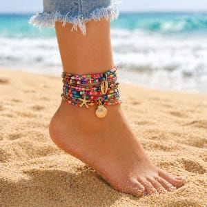 May include: A multi-strand beaded anklet with colorful beads, a gold starfish charm, and a gold seashell charm. The anklet is worn on a foot on a sandy beach with the ocean in the background.