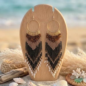 May include: A pair of long, dangling earrings featuring a gold-toned circular hoop at the top. The earrings are crafted with rows of small seed beads in a gradient pattern of metallic gold, brown, black, and white, arranged in a chevron fringe design.