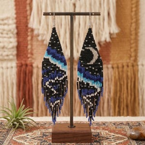 May include: A pair of long, teardrop-shaped beaded earrings featuring a night sky design. The black background is accented with a white crescent moon, scattered white stars, and horizontal bands of blue and white beads with fringe at the bottom.